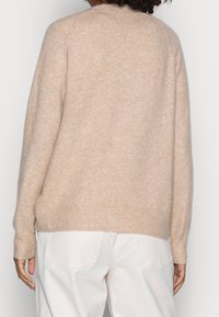 Vaaleanbeige neulepaita pyöreällä kaula-aukolla, ribbineuloksella helmassa ja pitkät hihat. Pehmeä pinta; takakuvassa näkyy minimaalinen suunnittelun yksityiskohdat.