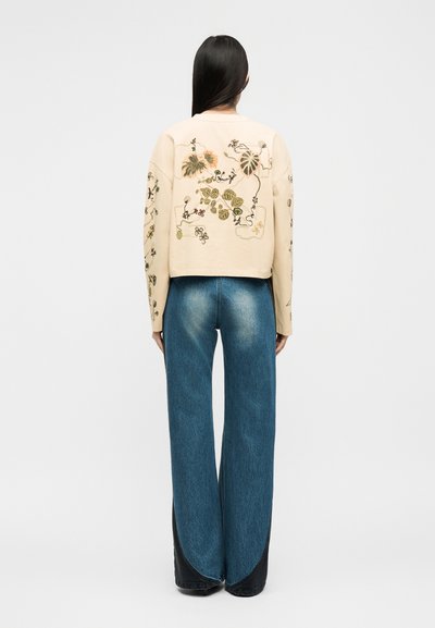 Γυναίκα με μακριά σκούρα μαλλιά φοράει μπεζ μπουφάν με κεντήματα λουλουδιών και φαρδιά μπλε τζιν με σκοτεινές καμπυλωτές πινελιές, στέκεται γυρισμένη προς τα πίσω.