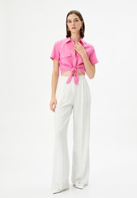Camisa corta rosa con detalle de nudo en la parte delantera, mangas cortas y dos bolsillos en el pecho, combinada con pantalones anchos de talle alto blancos.