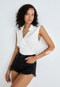 Chemise sans manches blanche avec un design enveloppant, un col et une texture lisse, associée à un short en denim noir taille haute avec un ourlet effiloché.
