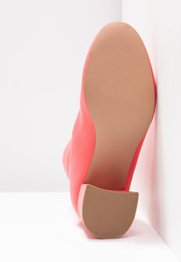 Chaussure à talon haut corail vif inclinée vers le haut, montrant une semelle lisse et un talon en caoutchouc texturé sur fond blanc.