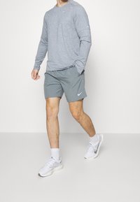 Harmaa pitkähihaiselta paita, harmaat shortsit Nike-logolla ja valkoiset urheilukengät. Asuste näyttää rentoa istuvuutta ja kevyitä materiaaleja.