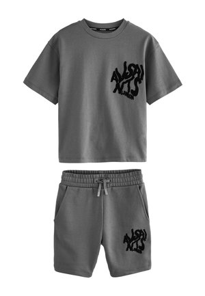 Blond chłopiec stoi, nosząc szary oversize’owy T-shirt i pasujące spodenki z czarnym nadrukiem, białe skarpetki i białe trampki na jednolitym tle.