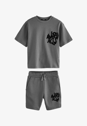 Blond jongetje staat in een grijs oversized T-shirt en bijpassende shorts met zwarte print, witte sokken en witte sneakers op een effen achtergrond.