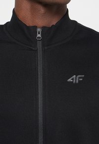 Chaqueta negra con cremallera y cuello alto, de tejido texturizado, y un sutil logotipo "4F" en una tonalidad más oscura en el lado izquierdo del pecho.