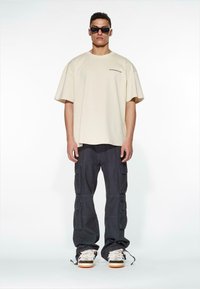 Beige oversized T-skjorte med korte ermer og logo; mørke cargo-bukser med lommer; hvite og grå sneakers; svarte solbriller.