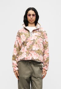 Γυναίκα που φοράει μπουφάν φλις με σχέδιο σε ροζ, καφέ και λευκό, με παντελόνι ελιάς και σκουρόχρωμα γυαλιά ηλίου, στέκεται μπροστά σε απλό φόντο.