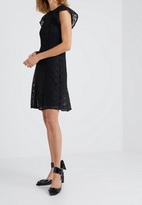 Abito nero in pizzo con maniche a volant e corpetto aderente, abbinato a scarpe con tacchi alti nere lucide decorate con accenti floreali.