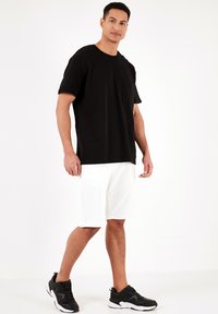 Sort T-shirt med korte ærmer og lomme, parret med hvide shorts til knæene og sorte træningssko, stående foran en ensfarvet baggrund.