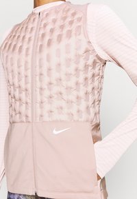 Jasnoróżowa kamizelka Nike z pikowanym górnym fragmentem, gładką dolną częścią i białym logo swoosh, noszona na długim rękawie z subtelnymi kropkowanymi paskami.