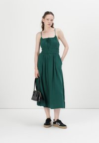 Grön, ärmlös maxiklänning med figursydd överdel, samlad midja och sidofickor. Kombinerad med svarta sneakers och en liten handväska.