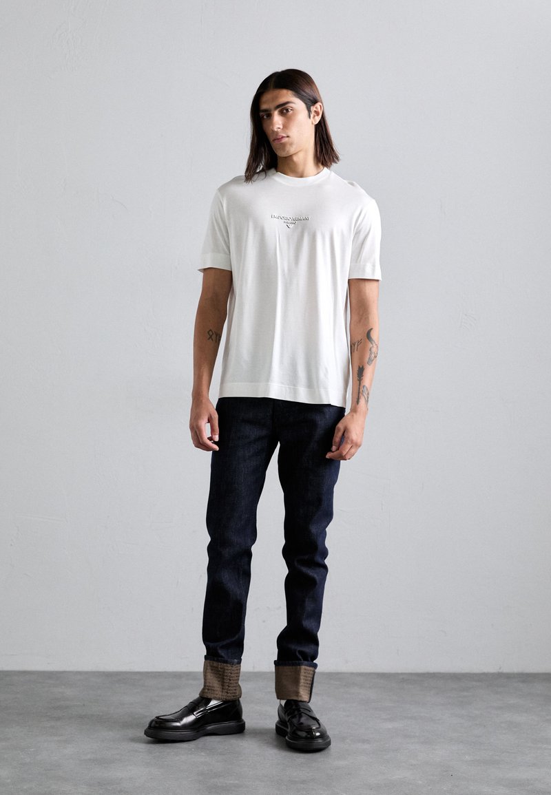 Man with shoulder-length hair wearing white short-sleeve shirt, dark rolled-up jeans, black shoes, standing against plain gray wall.
