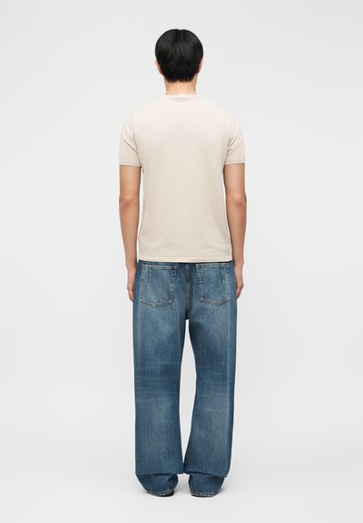 Άτομο με κοντά μαύρα μαλλιά που φοράει μπεζ πουκάμισο με κοντά μανίκια και φαρδιά μπλε τζιν, στέκεται με την πλάτη προς τα εμάς σε απλό φόντο.