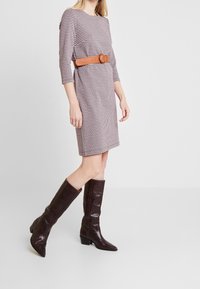 Femme portant une robe à motifs pied-de-poule longueur genou avec des manches trois-quarts, une ceinture beige et des bottes à talons hautes jusqu'au genou de couleur marron foncé.