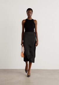 Top senza maniche nero, gonna midi in satin nera e tacchi a punta neri. La modella porta una piccola borsa a tracolla color pesco. Design semplice e elegante.