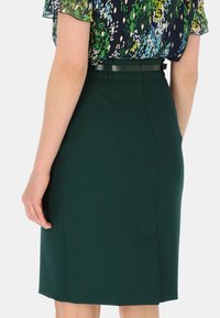 Zielona spódnica o gładkiej fakturze, do kolan, z rozcięciem z tyłu i subtelnym przeszyciem w talii, w połączeniu z wzorzystą bluzką.
