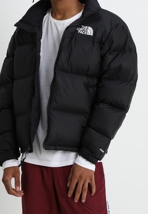 Person iført en sort dunjakke med The North Face-logo, hvid skjorte og bordeaux bukser, stående mod en enkel baggrund.