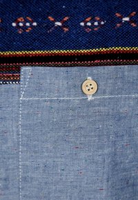 Tela azul claro con motas multicolores. Presenta un pequeño bolsillo en el pecho con un botón de madera. La parte superior muestra un ribete tejido con un patrón en colores oscuros.