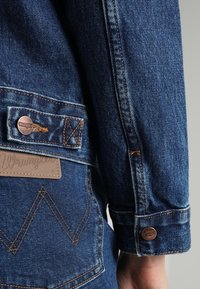 Detalj av denimjacka med blått tyg, kopparknappar, kontrastsöm och en läderlapp med präglad varumärkesbokstäver.