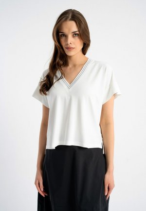 Kobieta z długimi brązowymi włosami, ubrana w biały top z krótkim rękawem i dekoltem w serek oraz czarną spódnicę, stojąca na jednolitym białym tle.