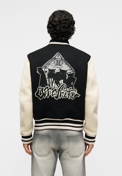 Άτομο που φοράει μαύρο και άσπρο varsity μπουφάν με γραφικό σχέδιο ενός σπιτιού και πέντε μαύρες γάτες, καθώς και στυλιζαρισμένο κείμενο στην πλάτη.