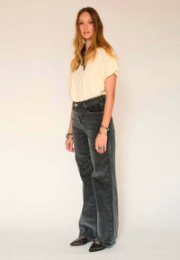 Top court jaune clair à manches courtes, pantalon large gris, chaussures noires à bout pointu avec des accents argentés, bracelets superposés aux poignets.