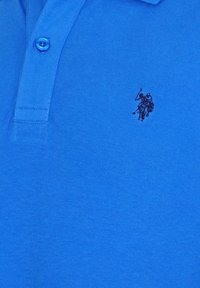 Blå pikétröja med en svart broderad logotyp av en polo-spelare på häst som svingar en klubba, nära knäppningen.