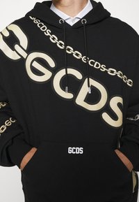 Felpa nera in morbido tessuto, con grandi lettere beige "GCDS" e motivo a catena. Include una tasca a marsupio e un cappuccio con lacci.