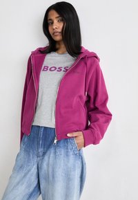Fuksija zip-hoodie s podstavljenom kapuljačom, nosi se preko sive majice s ljubičastim natpisom. Model kombinira s svijetloplavim, iznošenim širokim trapericama.