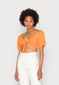Blusa laranja cropped com mangas bufantes, detalhe de laço na frente e bainha rendada. Usada com jeans brancos de cintura alta. Fundo simples.