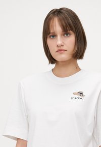 Tricou alb din bumbac, cu croi supradimensionat, prezentând un text brodat "BLAZING" și un grafic cu o pălărie de paie accentuată cu o panglică albastră.