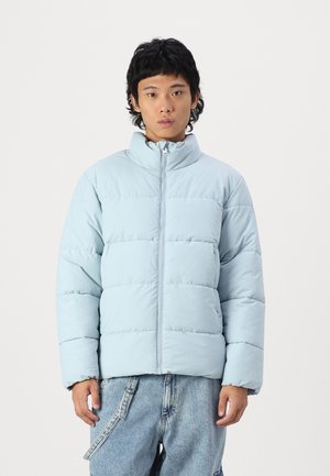 Jachetă puffer de culoare albastru deschis, cu guler înalt, quilt-ing orizontal, fermoar frontal și mâneci lungi, asortată cu pantaloni din denim.