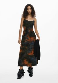 Donna con capelli ricci che indossa un vestito lungo senza maniche e con motivo, tiene in mano una giacca nera e indossa stivali in pelle neri e morbidi.