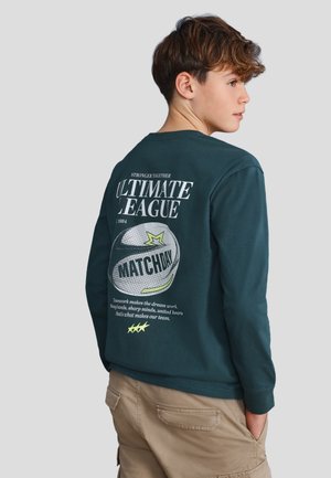 Chłopiec patrzący w bok, ubrany w ciemnozieloną koszulkę z długim rękawem z napisem "Ultimate League Matchday" i grafiką siatkówki na plecach.