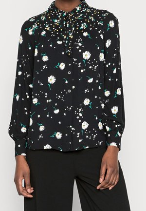 Blusa negra de manga larga con estampado de margaritas blancas y pequeñas estrellas, con botones en la parte delantera y detalle de lazo en el cuello, combinada con pantalones negros.