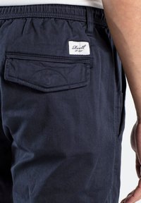 Pantalones de algodón azul marino con una cinturilla elástica, que cuentan con un bolsillo trasero con tapa y una etiqueta que dice "Reell est. 1997."