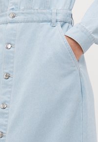 Svetlomodré denimové šaty so striebornými gombíkovými uzávermi, opaskom v páse a bočnými vreckami. Majú hladkú textúru a dlhé rukávy.