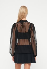 Blusa preta translúcida com mangas longas, ligeira renda no colarinho e detalhe de abertura nas costas. Usada sobre um top preto ajustado.