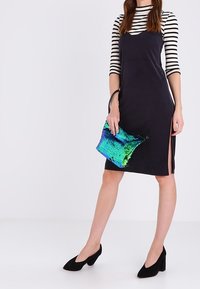 Abito nero aderente con spacco laterale, indossato sopra una maglia a righe bianco e nero. Tiene in mano una borsa di paillettes multicolore. Tacchi neri completano l'outfit.