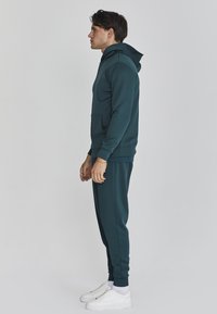 Sudadera azul petróleo con bolsillo canguro y puños acanalados, combinada con pantalones de chándal ajustados a juego. Zapatillas blancas completan el atuendo.