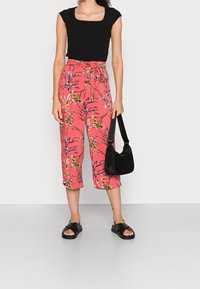 Zwarte fitted top, roze bloemenpatroon cropped broek met strik taille, draagt zwarte slide sandalen en houdt een zwarte handtas vast.