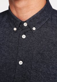 Close-up van een persoon die een donderblauwe overhemd met knopen en witte knopen draagt, waarbij een deel van de hals en het bovenste deel van de borst zichtbaar is.