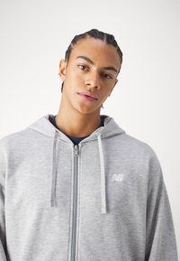 Grå lynlåshættetrøje med en blød tekstur, prydet med et hvidt logo på brystet og snore i hætten. Enkel, sporty design.
