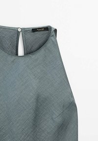 Débardeur sans manches de couleur gris clair, en tissu texturé, avec un col rond, un système de fermeture à boutons au dos et un détail de poche discret.