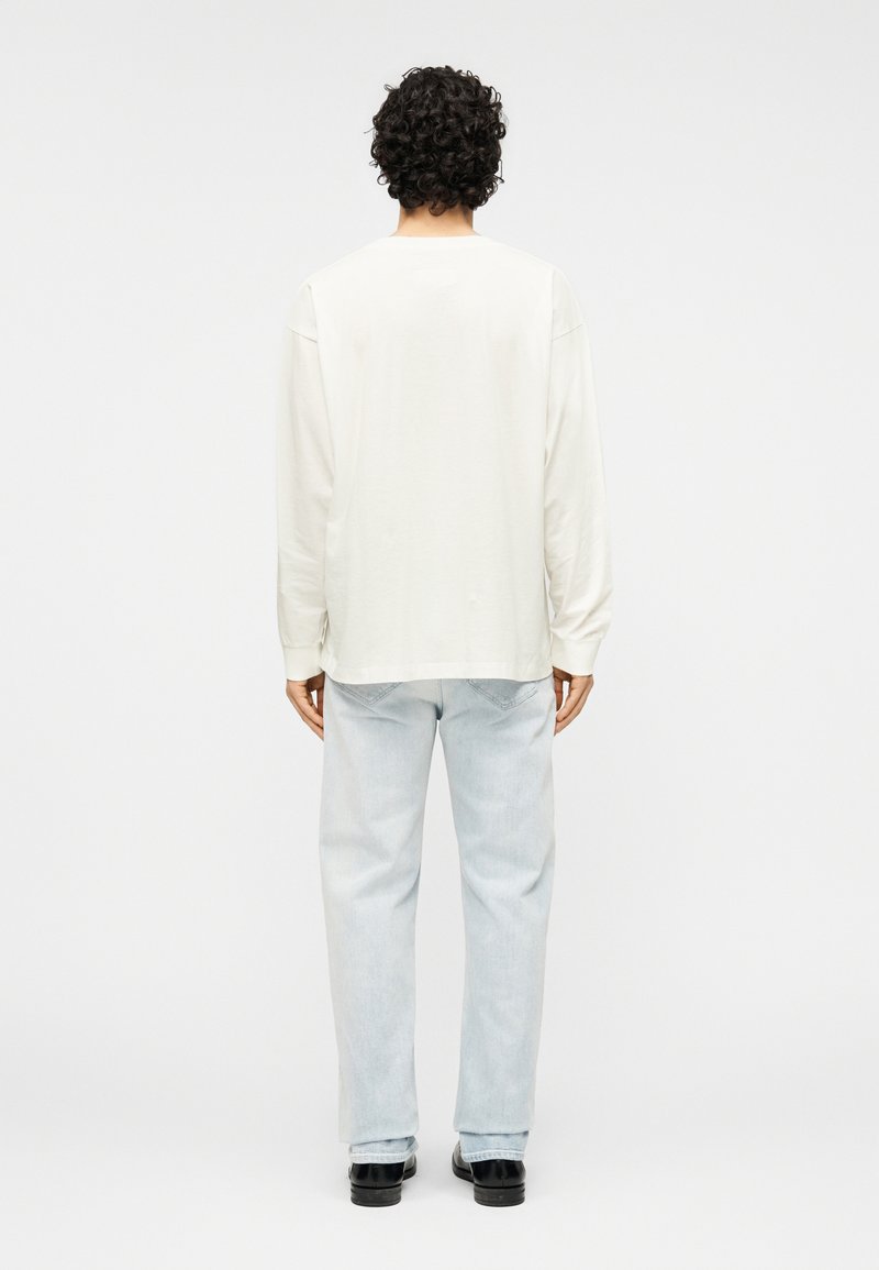 Chemise à manches longues de couleur blanche cassée avec une coupe décontractée, associée à un jean bleu clair. La chemise présente une texture simple et lisse.