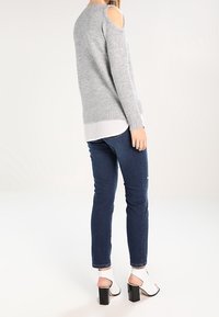 Grijze gebreide trui met uitsneden op de schouders, gecombineerd met donkerblauwe skinny jeans. Model draagt witte open teenhakken met blokhakken.