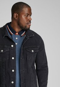 Black denim jacket with silver buttons and pockets, layered over a blue polo featuring a contrasting collar with stripes.