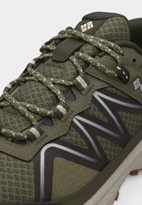 Αθλητικό παπούτσι από πλέγμα σε ελιά πράσινο με κορδόνια με σχέδιο, γωνιώδη μαύρο και ασημί μοτίβο και επενδυμένη γλώσσα σε λευκή σόλα.