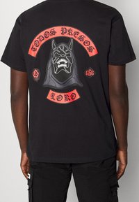 Černé bavlněné tričko s velkým potiskem na zádech: kostlivá postava, červené akcenty a text v stylizovaném písmu.