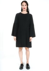 Crna haljina s zvonastim rukavima, okruglim izrezom, slobodnog kroja i do koljena. Uparena s crnim ravnim cipelama, postavljena na bijeloj pozadini.